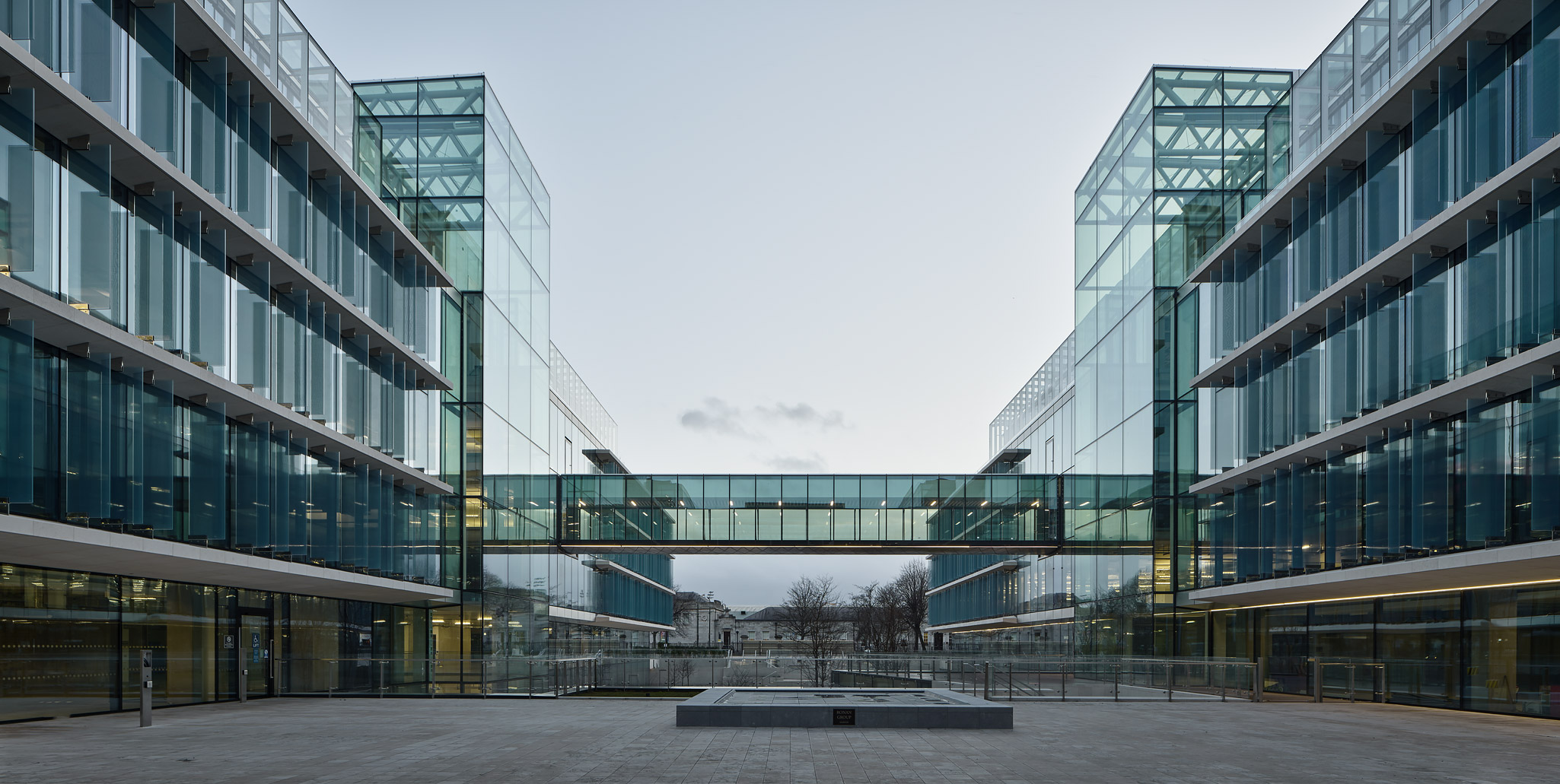 Interior courtyard view of Fibonacci Square office development by Johnny Ronan's Ronan Group in Ballsbridge Dublin, showing two symmetrical modern glass buildings connected by elevated walkway, with central plaza featuring Ronan Group plaque and views to historic RDS buildings beyond, located opposite Royal Dublin Society in Dublin 4